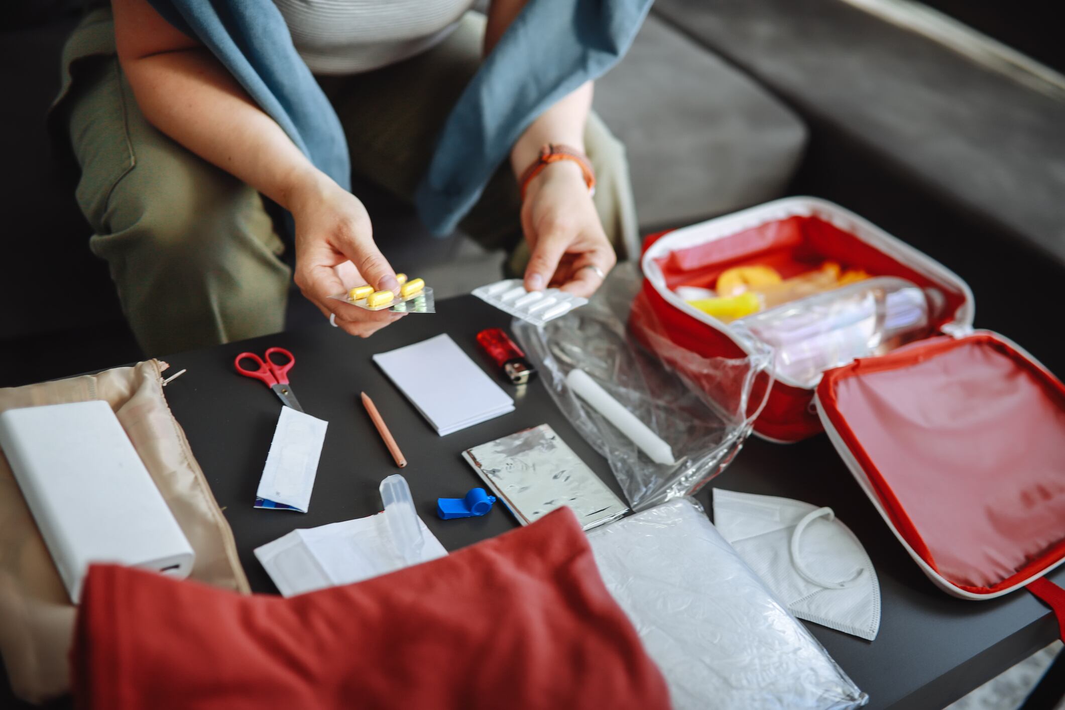 Plus-size woman in middle age checks medicines in a doomsday prepping kit at a table in a living room. Packing warm clothes, flashlight, power bank, medicines, water, and other emergency essentials for natural disasters.