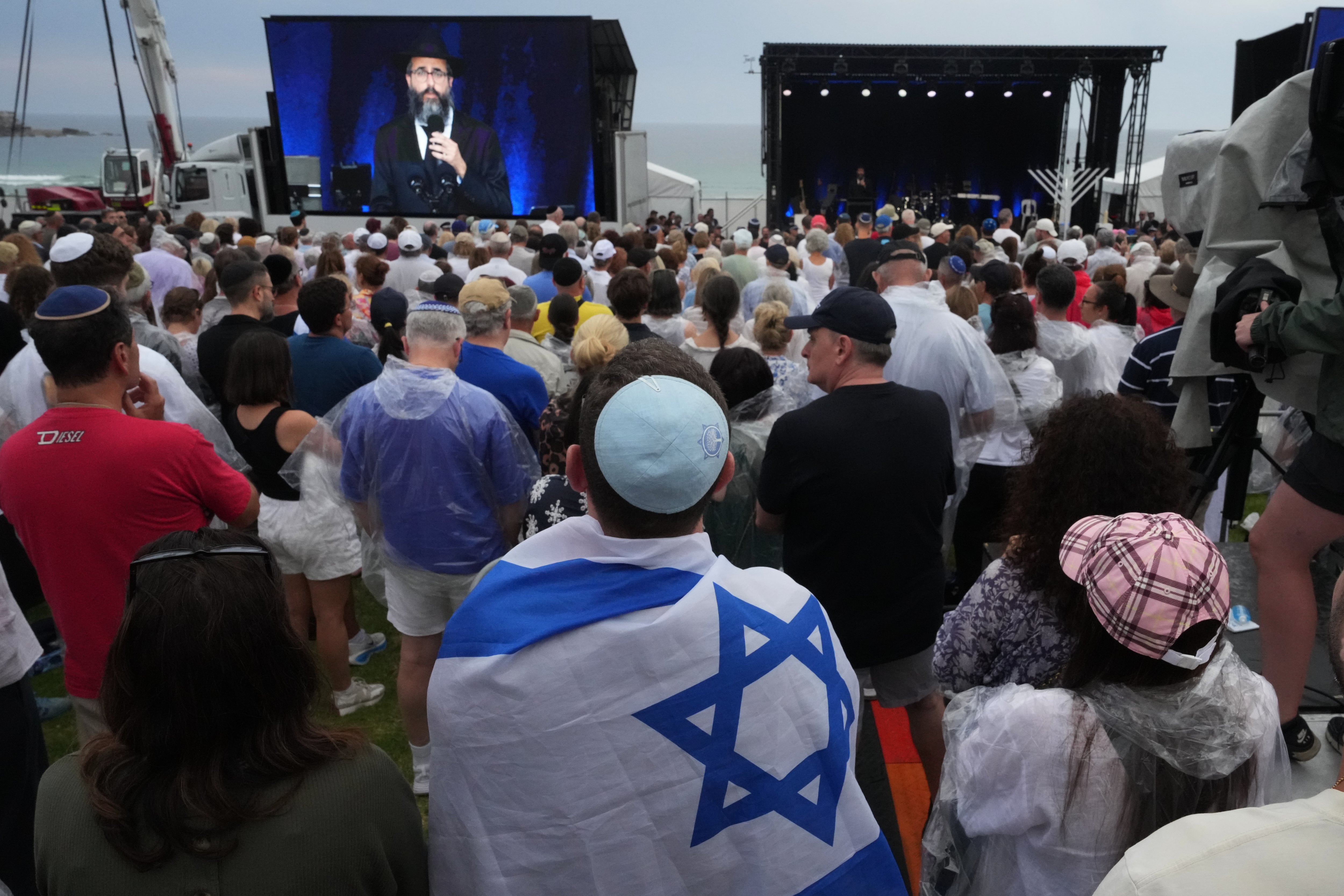 Una persona con la bandera de Israel y otras personas asisten a una ceremonia para conmemorar el Día Nacional de Reflexión por las víctimas y los sobrevivientes, en la playa de Bondi, Sídney.