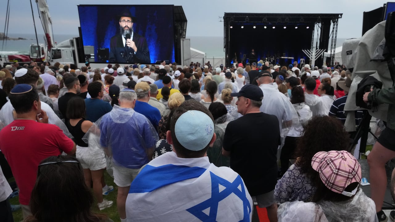 Una persona con la bandera de Israel y otras personas asisten a una ceremonia para conmemorar el Día Nacional de Reflexión por las víctimas y los sobrevivientes, en la playa de Bondi, Sídney.