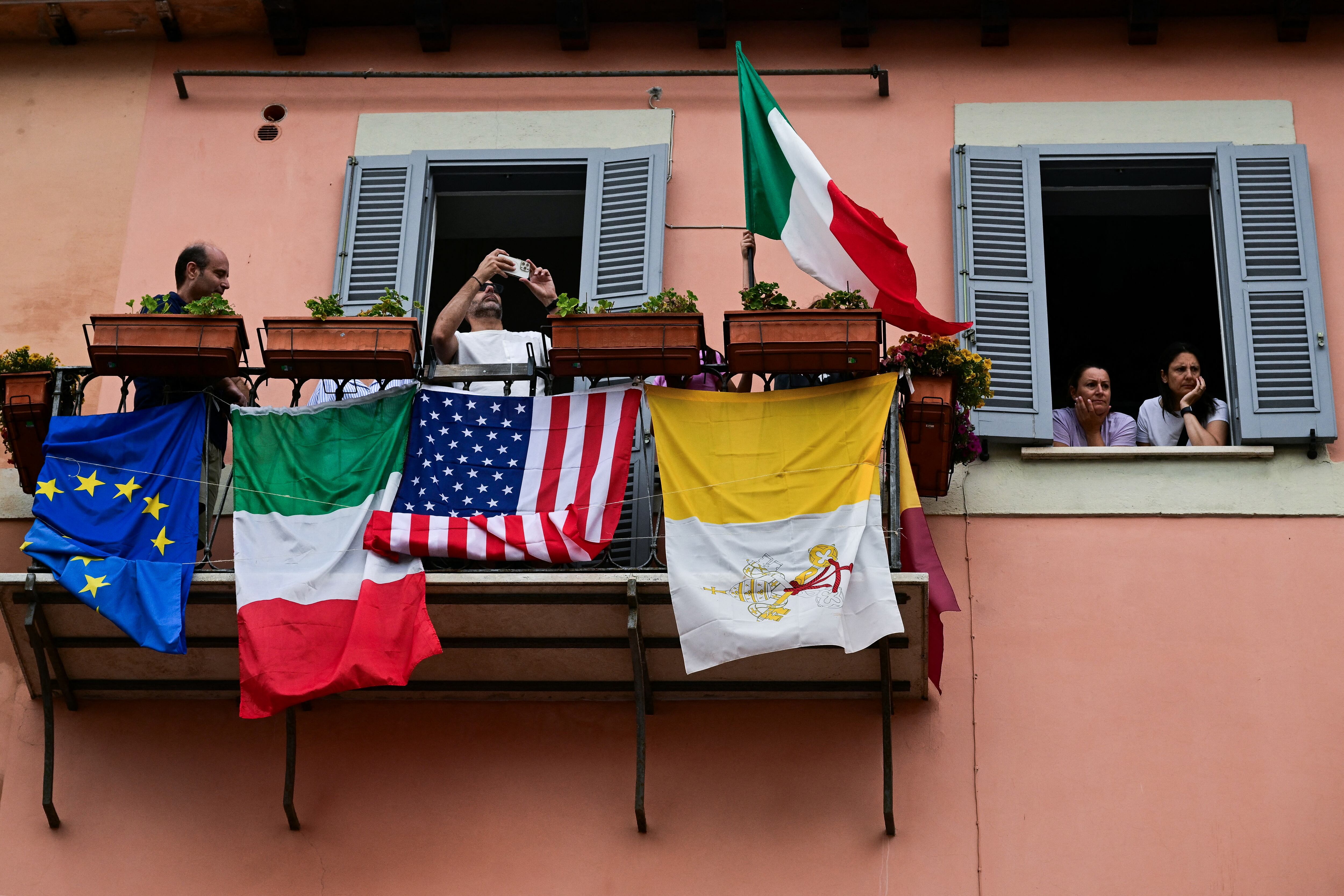 La gente con la bandera de la Unión Europea, Italia, Estados Unidos y el Vaticano escucha al Papa León XIV dirigiéndose a la multitud para la oración del Ángelus en la Piazza della Liberta (Plaza de la Libertad) frente al Palacio Apostólico (Palacio Apostólico) en la finca papal de verano en Castel Gandolfo, a 40 km al sureste de Roma, el 13 de julio de 2025. El recién elegido Papa León XIV, toma un descanso de verano del 6 al 20 de julio en el palacio papal de Castel Gandolfo, en las afueras de Roma, una antigua residencia de campo para pontífices que Francisco se negó a utilizar. (Foto de Tiziana FABI/PISCINA/AFP)
