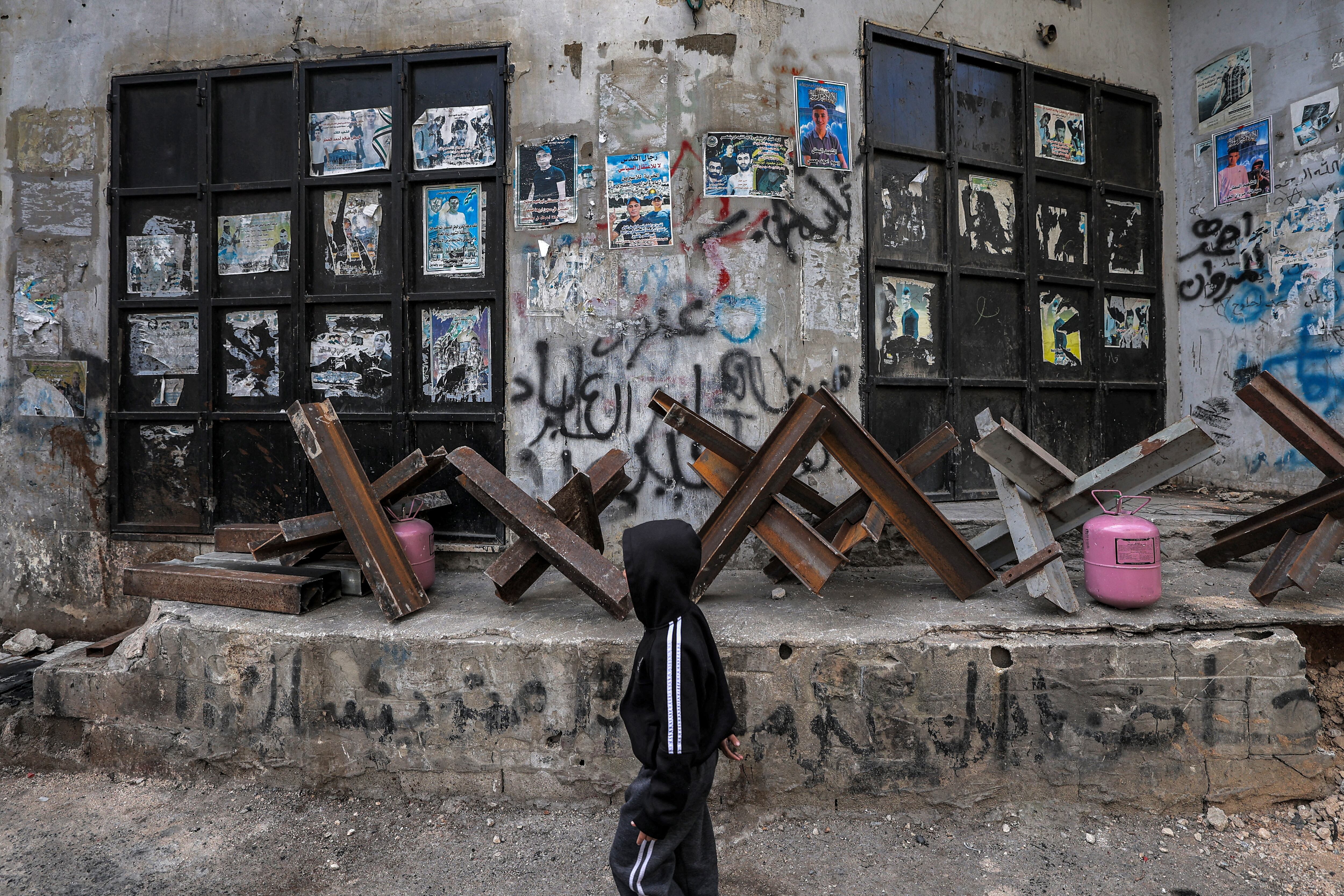 Un niño pasa junto a obstáculos de acero antivehículo colocados fuera de tiendas cerradas en el campamento de refugiados palestinos de Yenin en la Cisjordania ocupada el 8 de marzo de 2023 durante una huelga general palestina convocada en protesta contra la incursión del ejército israelí allí el día anterior. (Foto de JAAFAR ASHTIYEH / AFP)