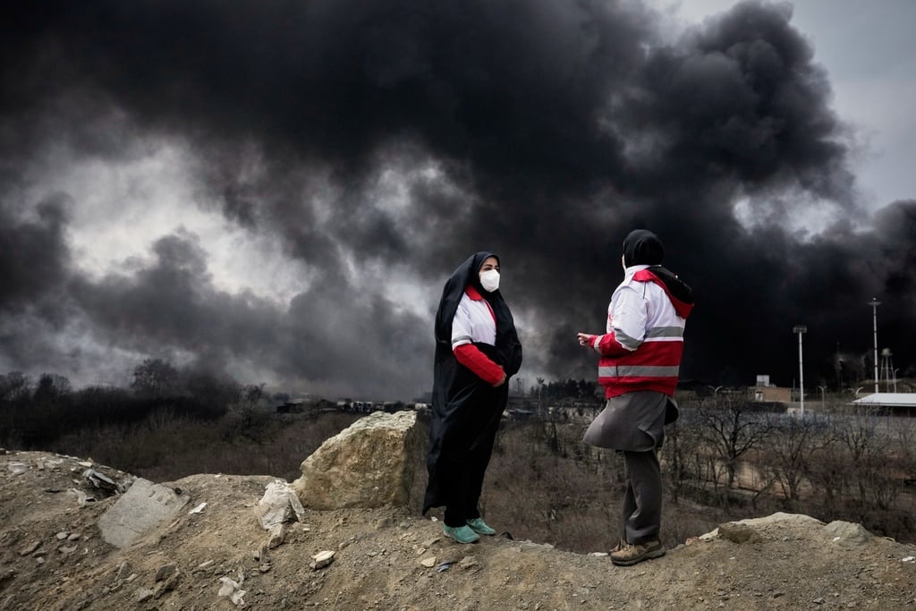 Dos mujeres de la Sociedad de la Luna Roja se ven de pie con una densa humareda provocada por un ataque el sábado por la noche de la campaña estadounidense e israelí contra una instalación petrolera, en Teherán, Irán, el domino 8 de marzo de 2026. (AP Foto/Vahid Salemi)