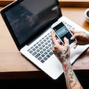 A close up shot of a young tattooed woman's hands checking her smart phone in a café while working on her Laptop. The room is bright and there' a cup of coffee in front of her.