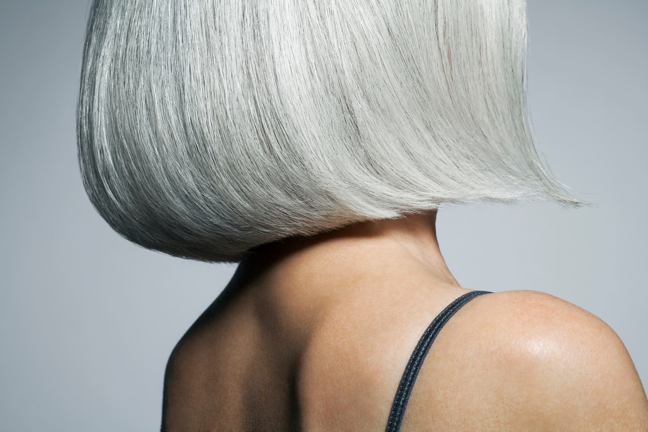 Mature woman with silvery, grey hair styled in a chin length bob in front of a grey background wearing a spaghetti strap tank top turning away from camera, cropped.
