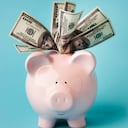 "Una alcancía rosa sonriente llena de billetes de 100 dólares, sobre fondo azul con espacio para copiar. También te puede interesar:"