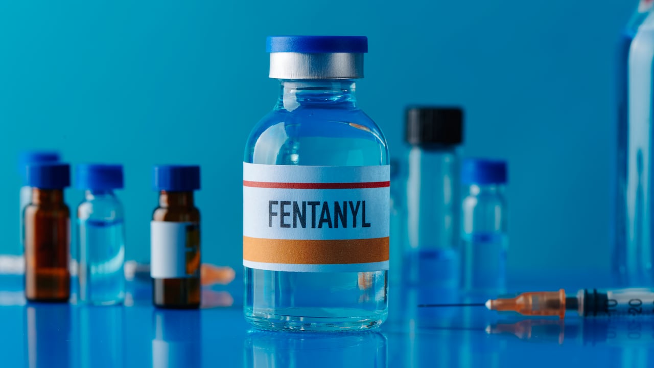 Primer plano de un vial simulado de fentanilo sobre una mesa azul junto a una jeringa y otros viales diferentes.