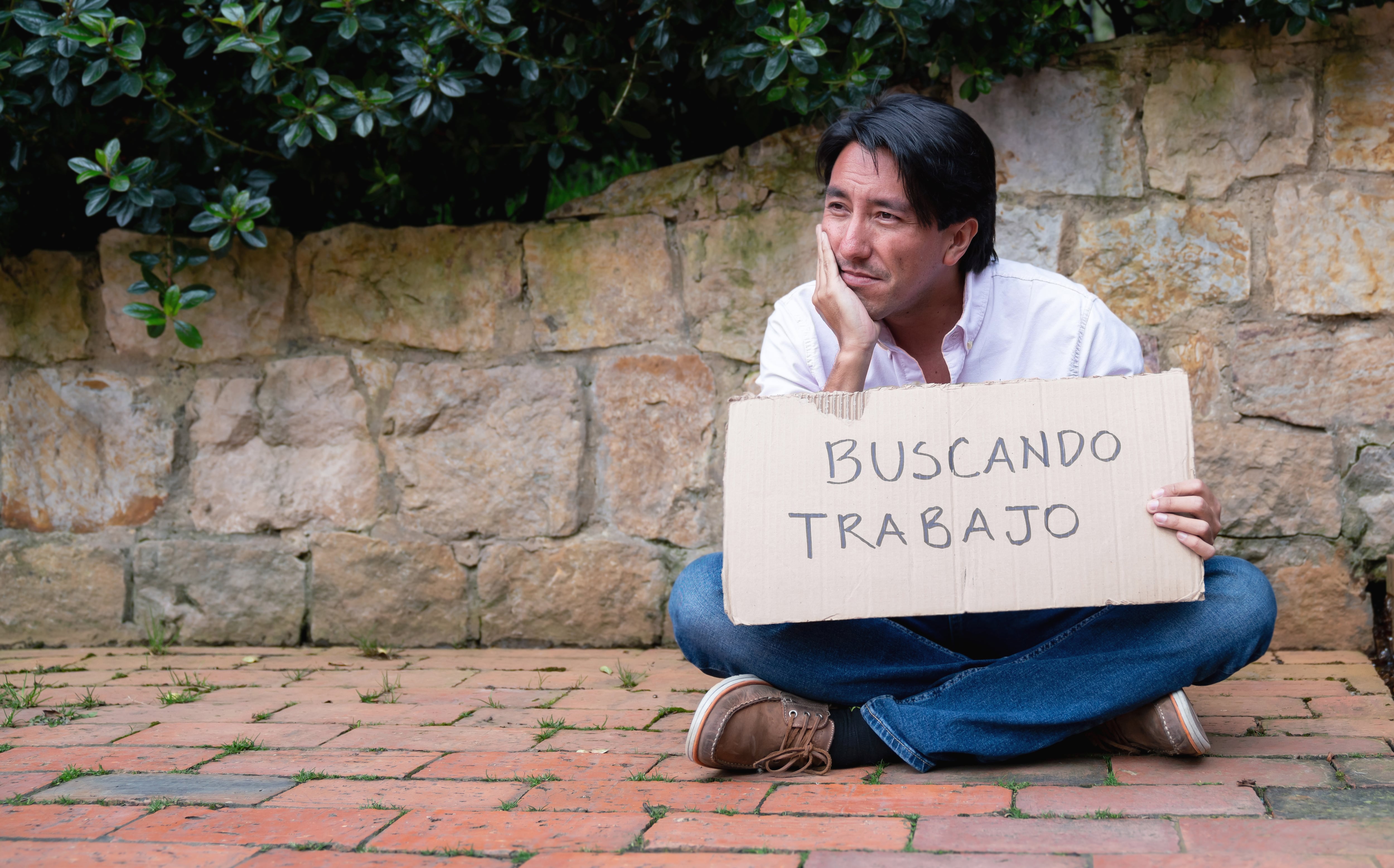 Man sitting on the street with a cardboard sign looking for a job written in Spanish