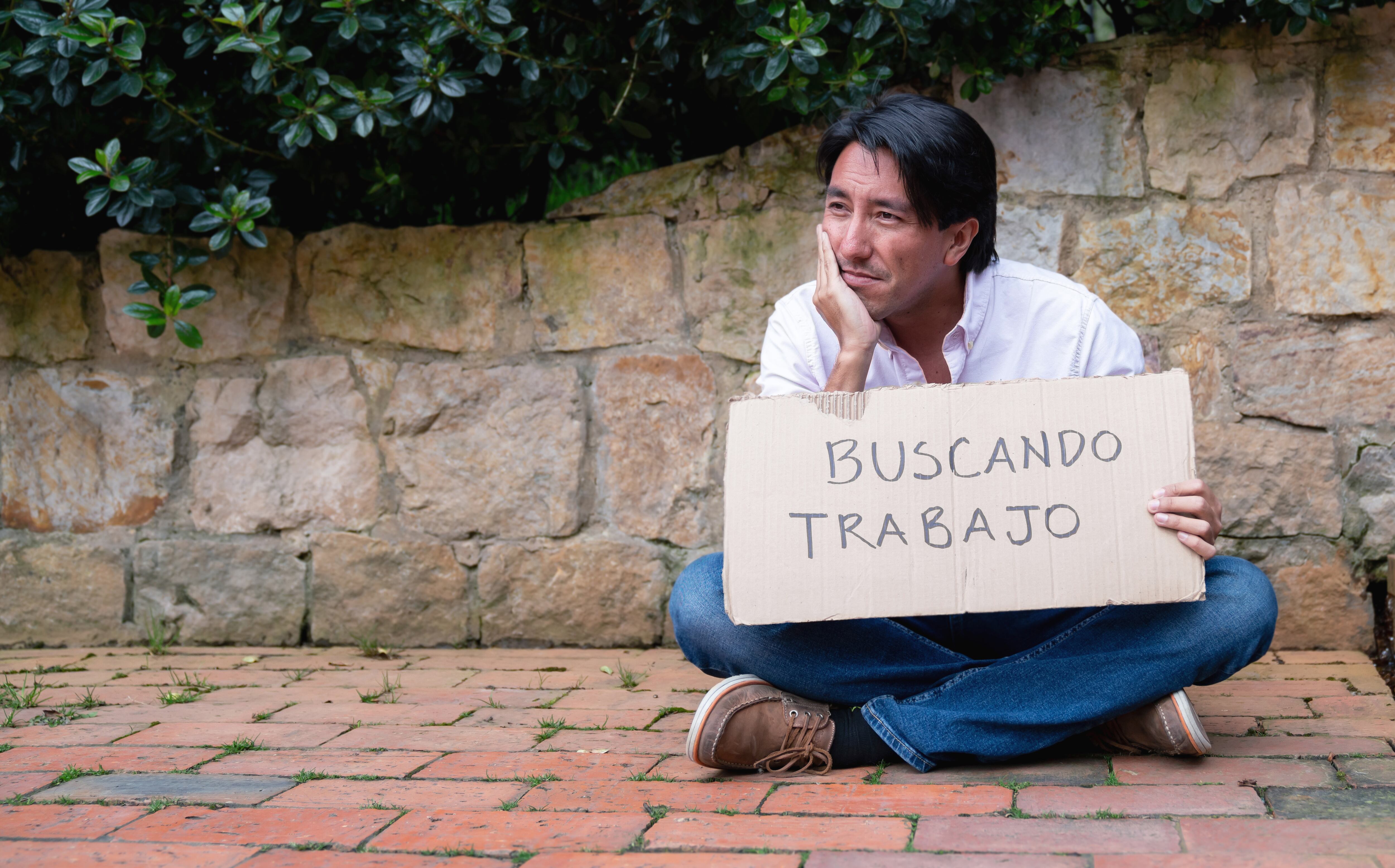 Man sitting on the street with a cardboard sign looking for a job written in Spanish