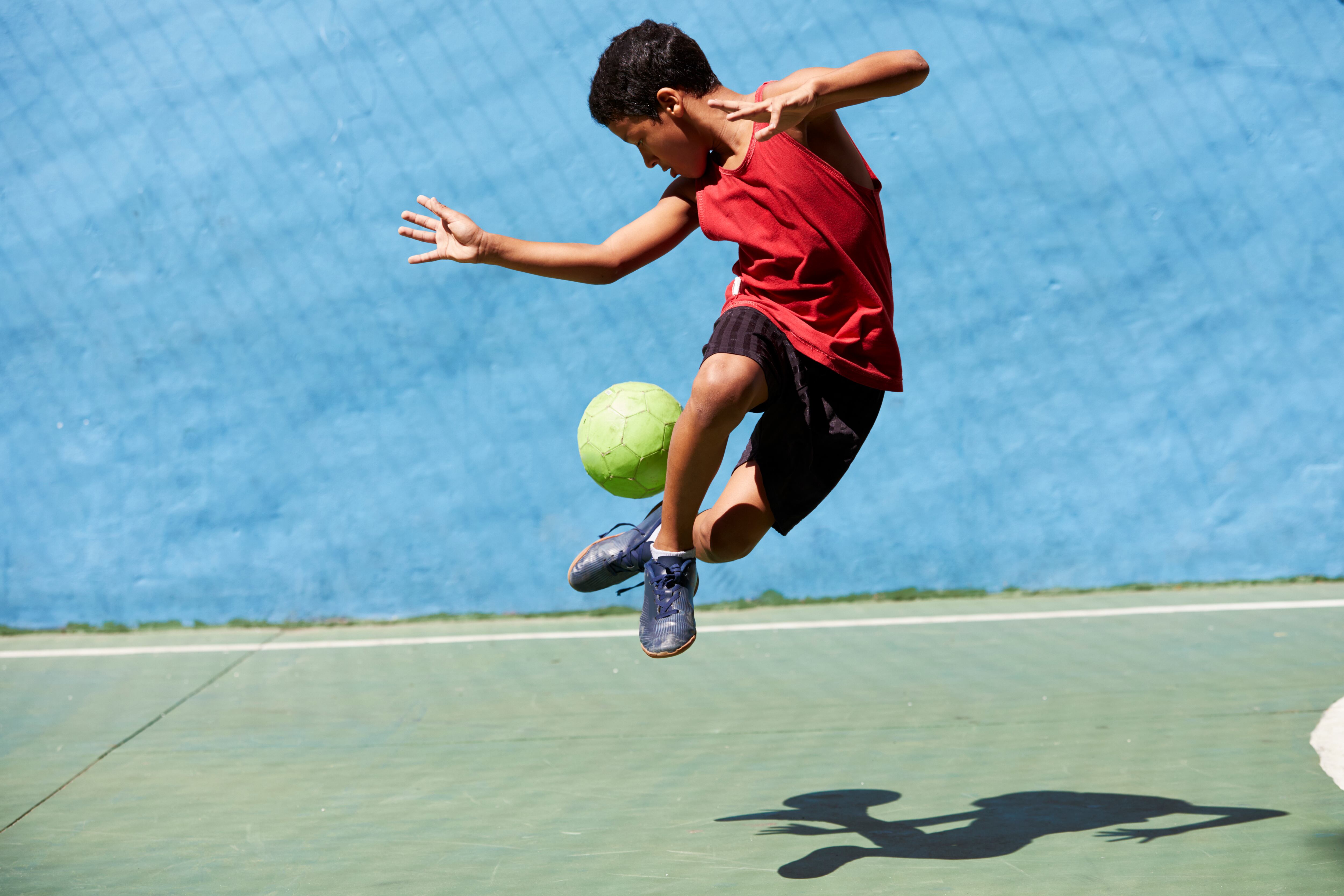 Joven brasileño jugando al fútbol en una cancha de fútbol de colores brillantes en una favela de Río. Lleva un chaleco rojo, pantalones cortos negros y botas azules. La bola es de color verde brillante.