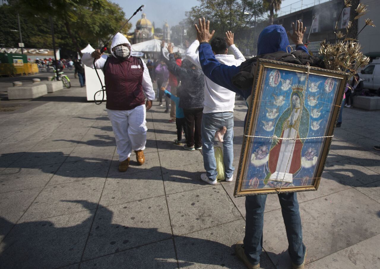 Un trabajador sanitario desinfecta a peregrinos en su camino hacia la Basílica de la Virgen de Guadalupe para frenar la propagación del coronavirus, mientras Alejandro Castillo carga con una imagen de la virgen a su espalda, en la Ciudad de México, el 9 de diciembre de 2020. Castillo partió de su casa en San Gregorio Atzompa, en el estado de Puebla, junto a su familia para peregrinar hasta la basílica el 6 de diciembre, y llegó al templo un día antes de que cerrase por la pandemia de la covid-19 . (AP Foto/Marco Ugarte)