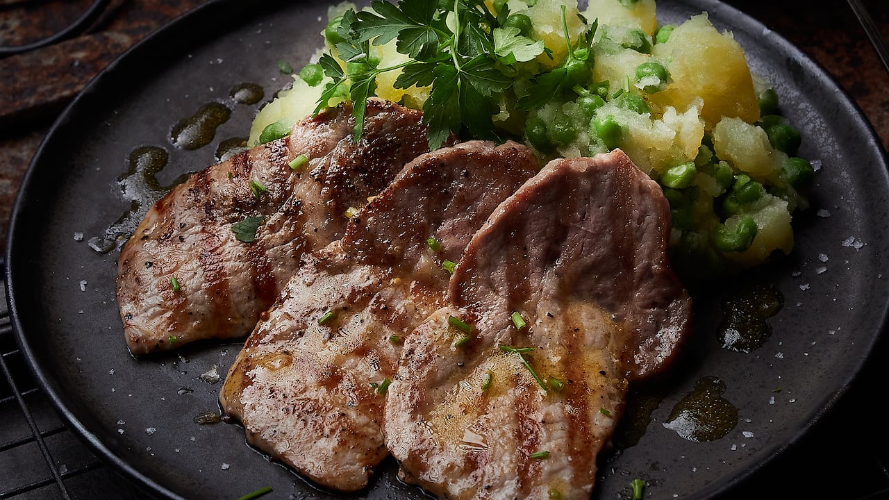 three grilled pork tenderloin steaks on black plate with side of boiled potatoes, peas and parsley. dressing sauce in a bowl on the side
