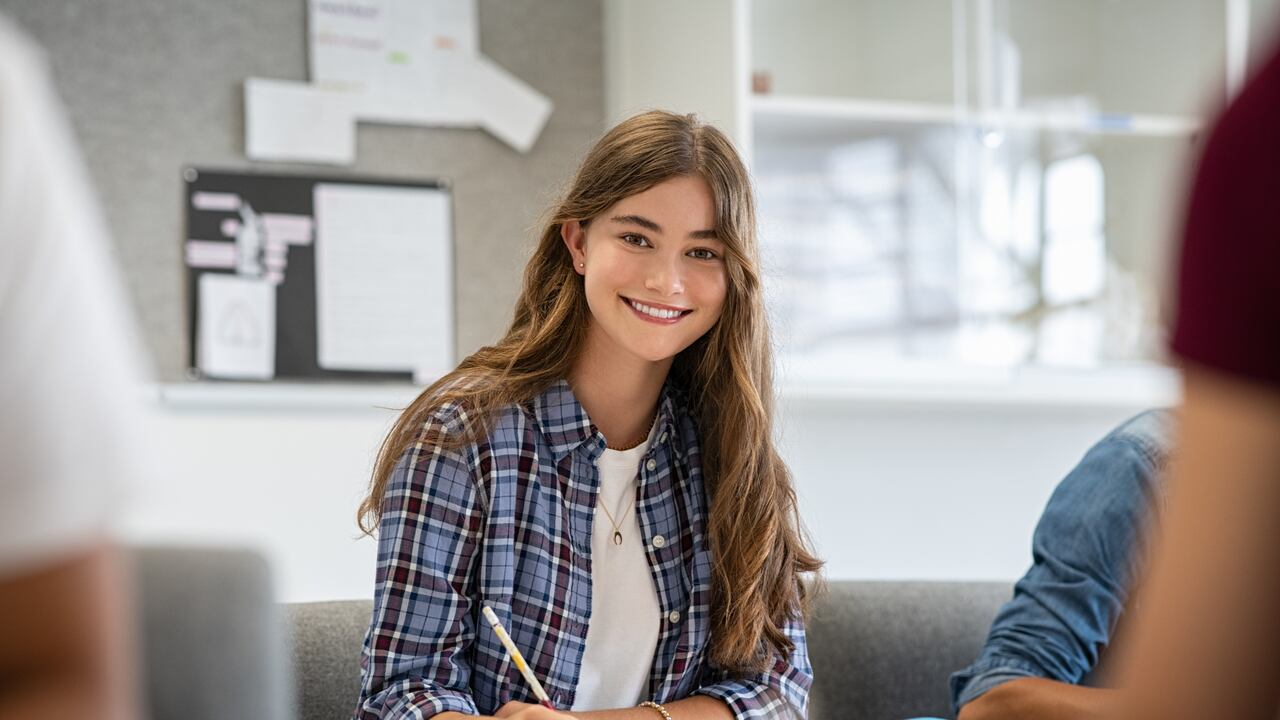 Beautiful university student smiling while studying for exams in classroom. Pretty woman sitting in classroom full of students during class. Portrait of happy young woman writing notes and looking at camera.