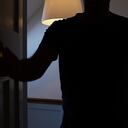 Silhouette of a man opening a door to a interior dark room, possibly a bedroom. He is an unrecognisable figure outlined be the landing light behind him.