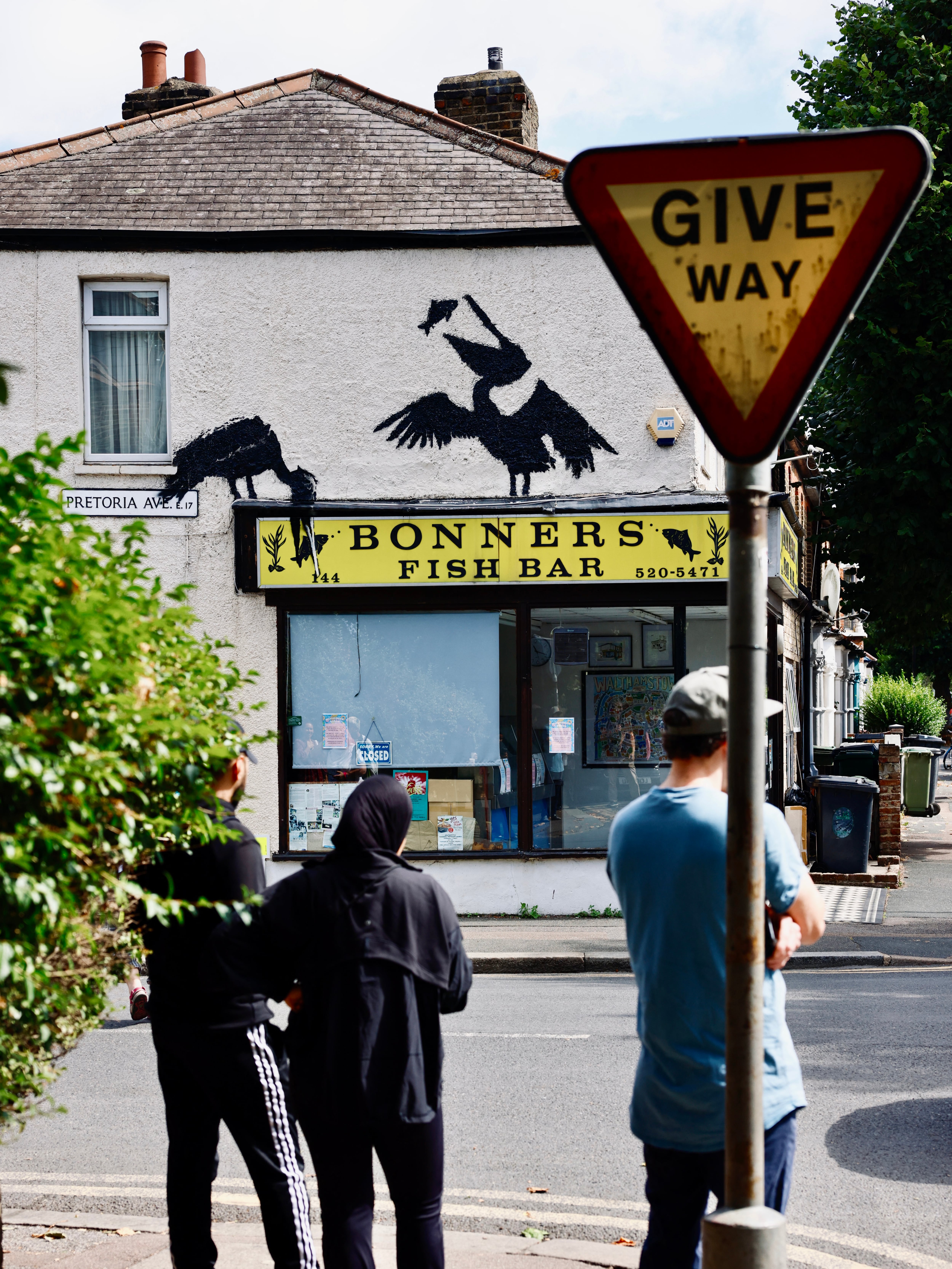 People gather to look at an artwork by street artist Banksy, the fifth to be released this week, depicting two pelicans catching fish, painted on top of a fish and chips shop in Walthamstow, northeast London, on August 9, 2024. The artist confirmed the work to be theirs after posting an image of it on the social media app Instagram. (Photo by BENJAMIN CREMEL / AFP) / RESTRICTED TO EDITORIAL USE - MANDATORY MENTION OF THE ARTIST UPON PUBLICATION - TO ILLUSTRATE THE EVENT AS SPECIFIED IN THE CAPTION