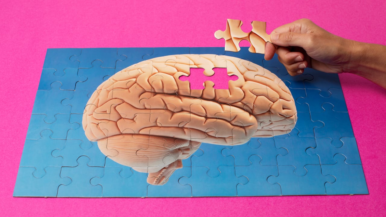 Mano de mujer a punto de completar el rompecabezas del modelo de cerebro humano sobre fondo azul y superficie magenta