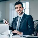 Foto de un joven empresario sentado en el escritorio en la oficina moderna y mirando a la cámara. Retrato de un apuesto hombre de negocios trabajando en equipo portátil y usando teléfonos inteligentes en la oficina.