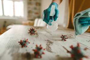 Mature woman cleaning and disinfects table in home office with a disinfectant and a rag. On the table is a model of a corona virus.