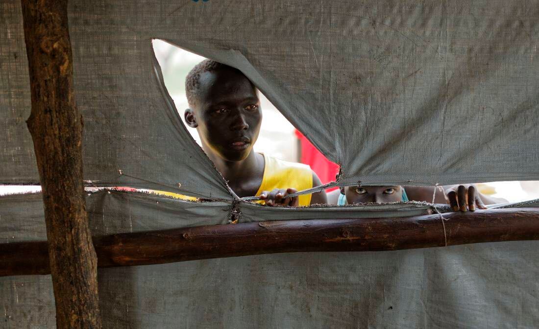Los refugiados del sur de Sudán miran por el lado desgarrado de una tienda de distribución de alimentos para ver cuándo empezarán a servir un almuerzo de puré de maíz y frijoles en el centro de recepción de Imvepi (Foto de AP / Ben Curtis)