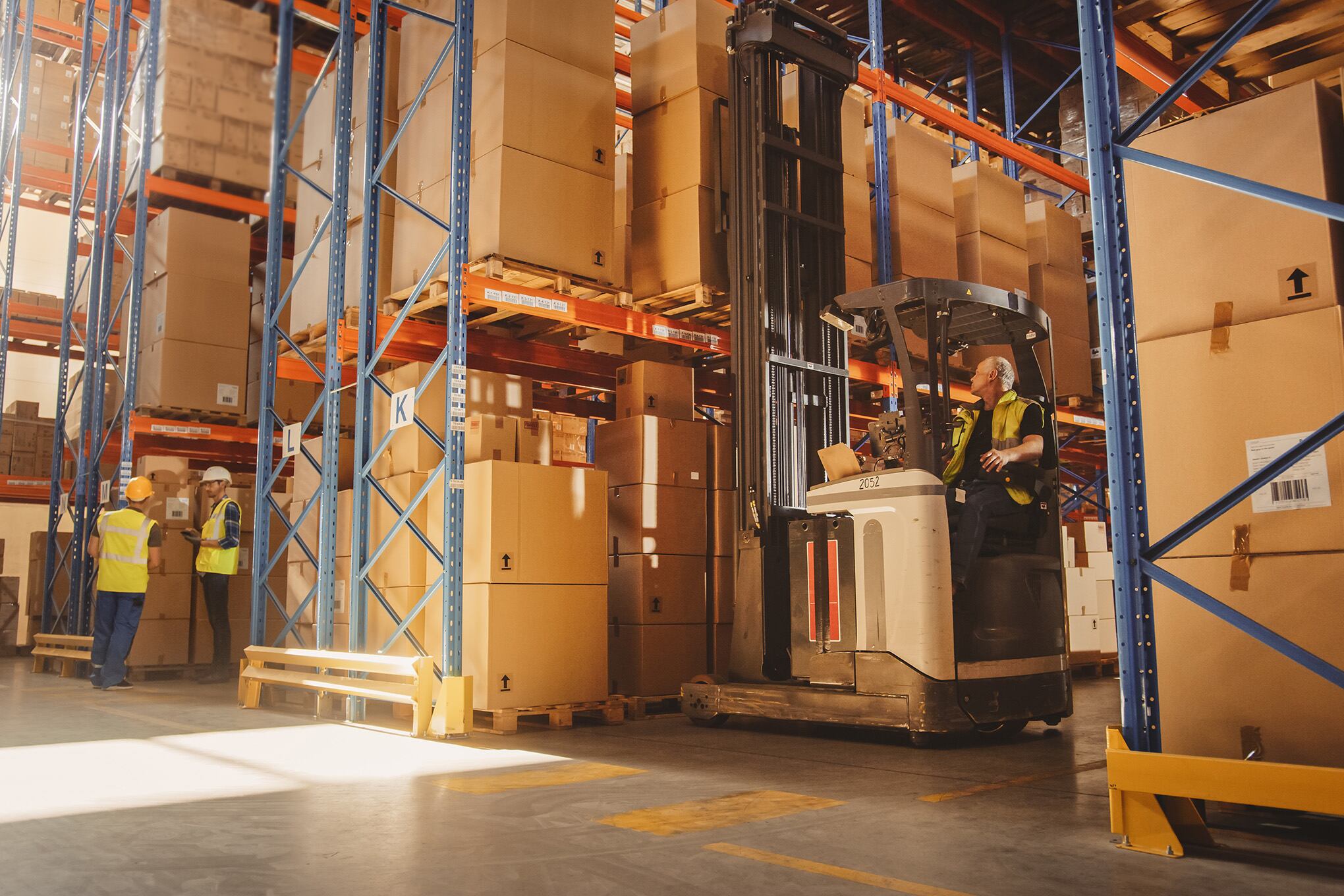 Almacén al por menor lleno de estantes con productos: el operador de carretillas elevadoras eléctricas levanta la paleta con la caja de cartón en un estante. Personas que trabajan, escanean productos, utilizando camiones en el centro de entrega de logística