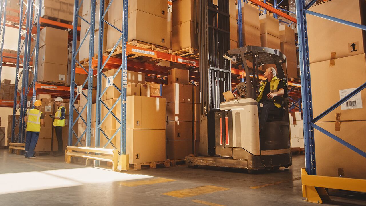 Almacén al por menor lleno de estantes con productos: el operador de carretillas elevadoras eléctricas levanta la paleta con la caja de cartón en un estante. Personas que trabajan, escanean productos, utilizando camiones en el centro de entrega de logística