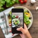 Blogger de comida usando un teléfono inteligente tomando fotos de una hermosa mezcla de ensalada verde fresca en una mesa de madera para compartir en las redes sociales
