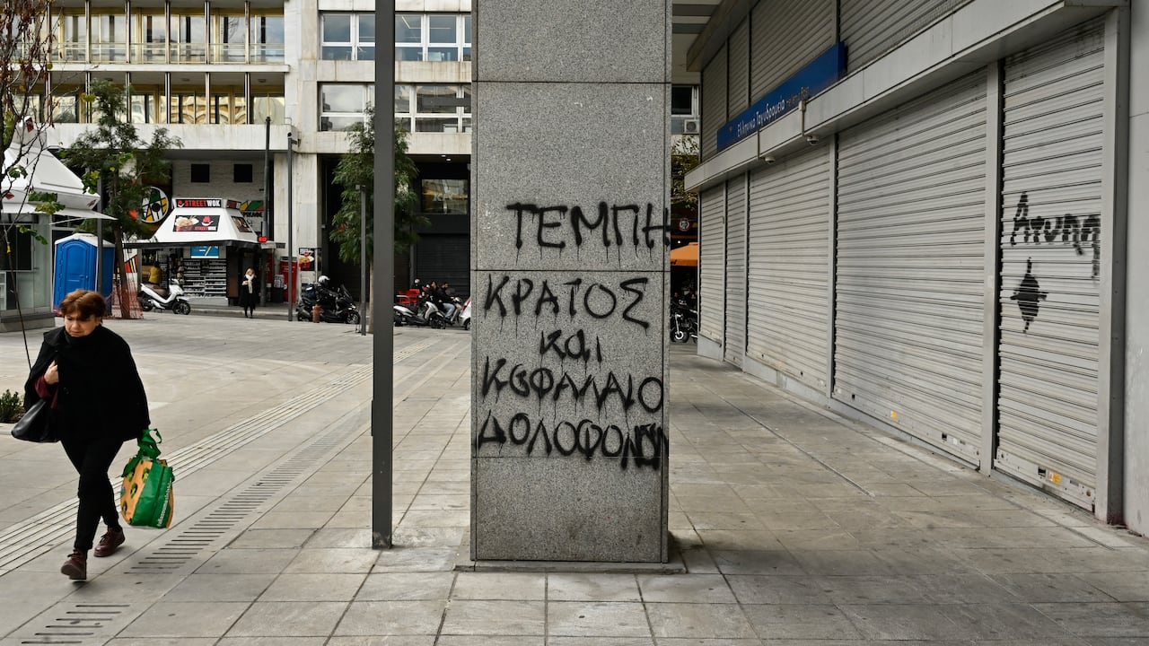 A woman walks by a graffiti reading "Tempi-state and capital murder" in front of closed postal service Hellenic Post, reffering to the deadly train crash, during a 24-hour strike in Athens on March 16, 2023. - Greek unions began a 24-hour walkout with demonstrations planned in major cities to voice outrage over last month's train disaster, which claimed 57 lives. (Photo by Louisa GOULIAMAKI / AFP)