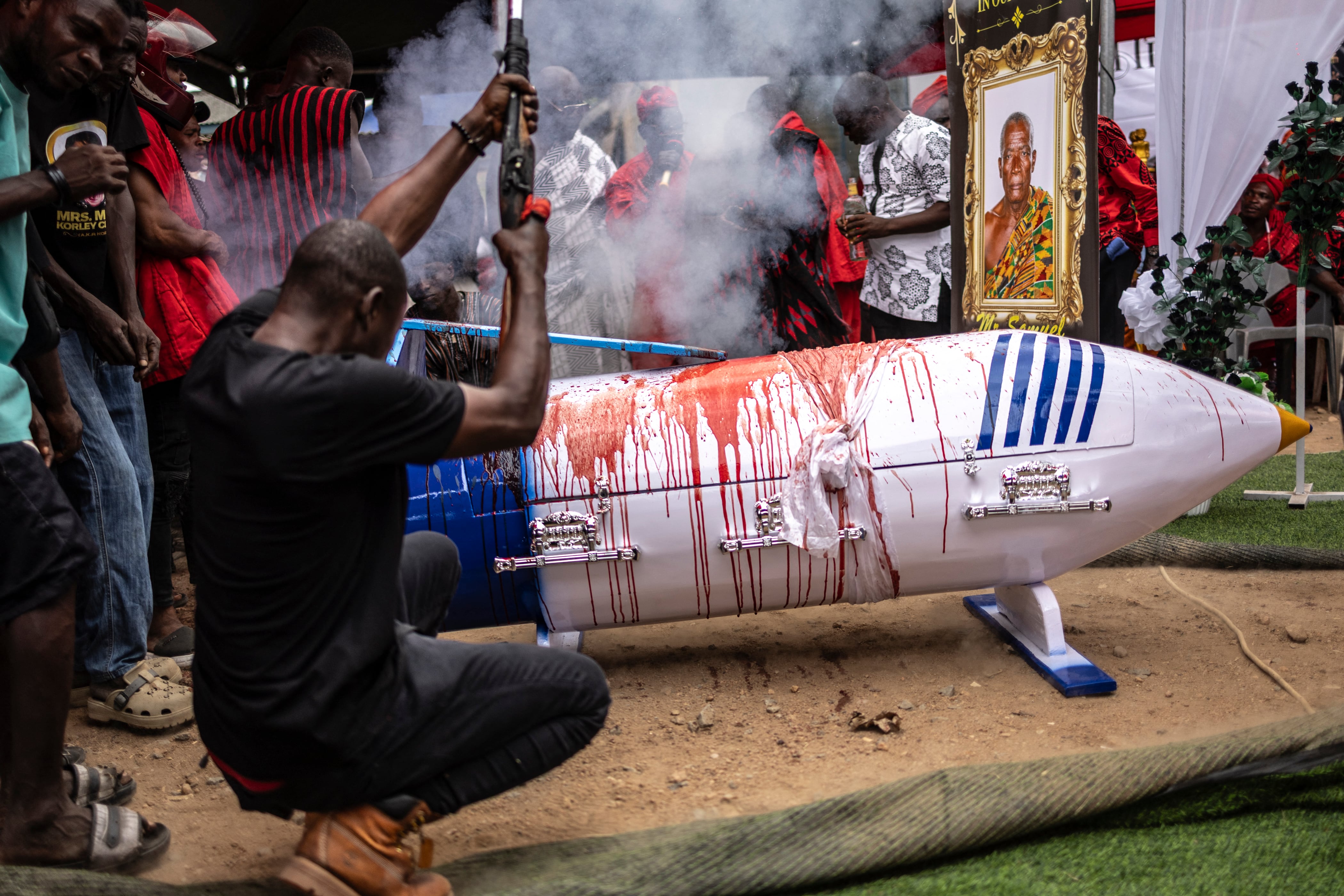 Un hombre dispara un arma de fuego tradicional junto al ataúd ensangrentado con forma de pluma del difunto profesor ghanés Samuel Commey Tetteh, después de que se sacrificara una oveja sobre él como parte de los rituales tradicionales durante su ceremonia fúnebre en Paanor, Ghana, el 15 de agosto de 2026.
Foto: OLYMPIA DE MAISMONT / AFP.
