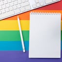 Bloc de notas, bolígrafo blanco, taza de café y teclado sobre fondo despojado de colores de la bandera del arco iris que simbolizan el movimiento LGBT.