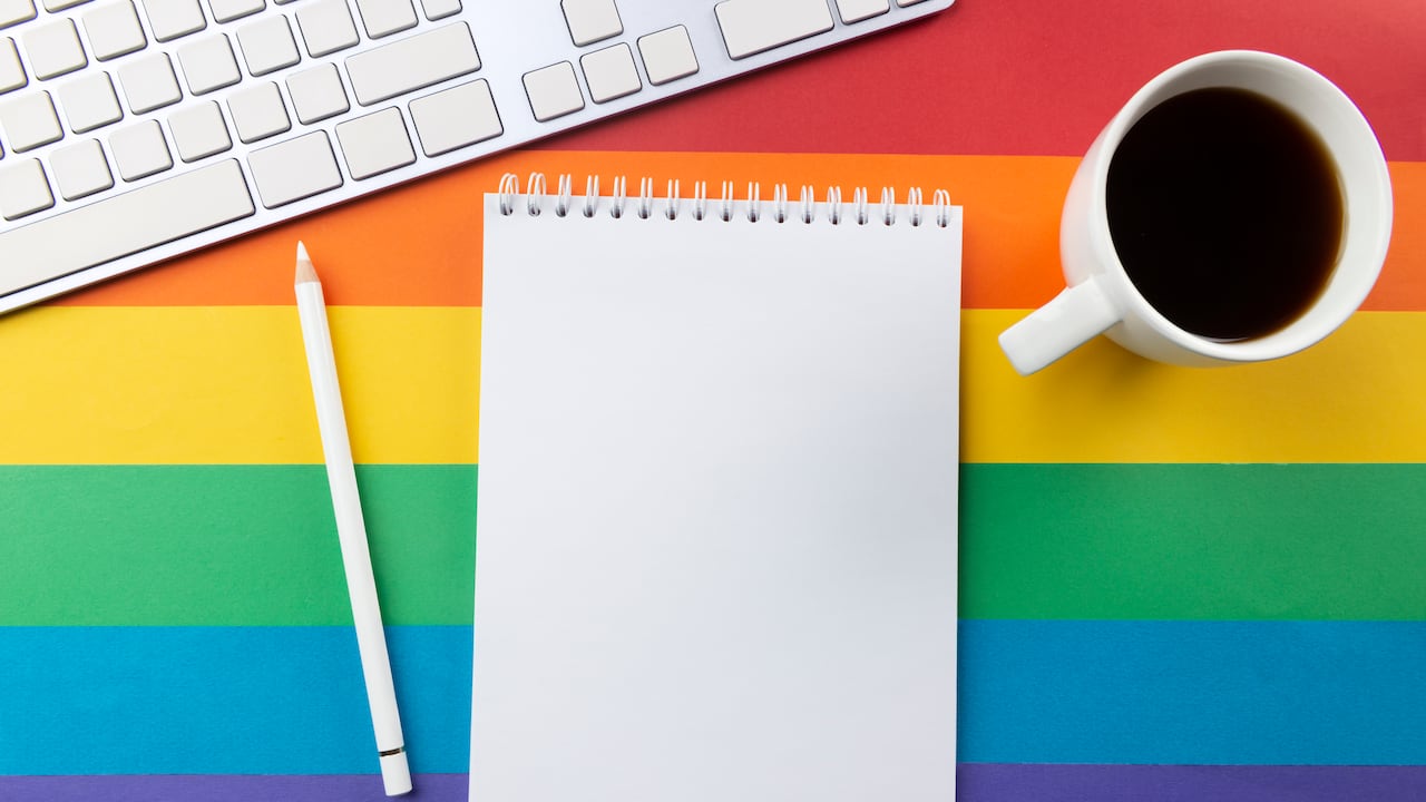 Bloc de notas, bolígrafo blanco, taza de café y teclado sobre fondo despojado de colores de la bandera del arco iris que simbolizan el movimiento LGBT.