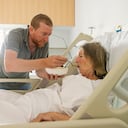 A woman (in her 70s) lies in a hospital bed having suffered a stroke. Her son (in his 40s) feeds her pureed food