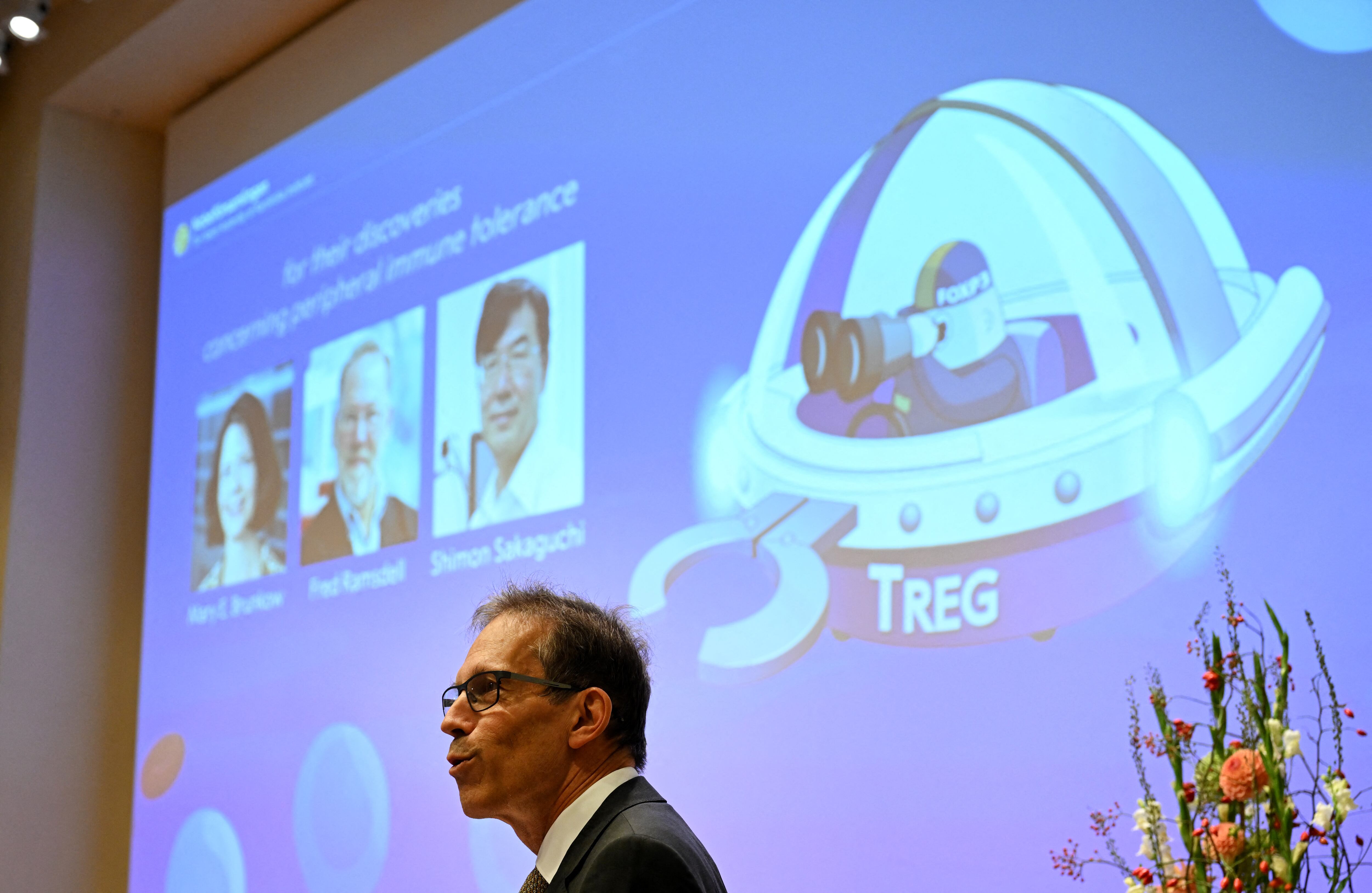 El secretario general del Comité Nobel, Thomas Perlmann  se dirige a la prensa frente a una pantalla con los retratos de Mary E. Brunkow, Fred Ramsdell y Shimon Sakaguchi.