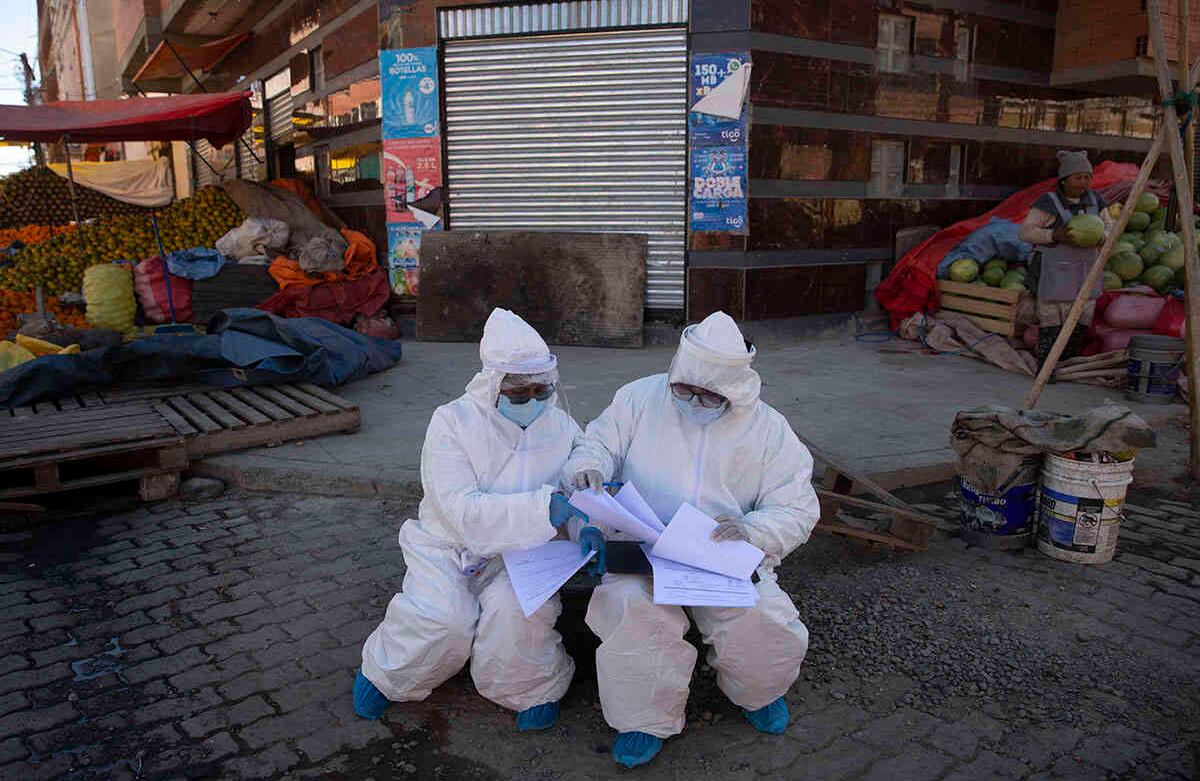 Los trabajadores de la salud vestidos con equipo de protección completo organizan sus documentos de datos que han recopilado durante una nueva prueba de coronavirus de casa en casa, rodeada por un mercado de productos en el barrio de Villa Dolores de El Alto, Bolivia, el sábado 18 de julio de 2020. (Foto AP / Juan Karita)