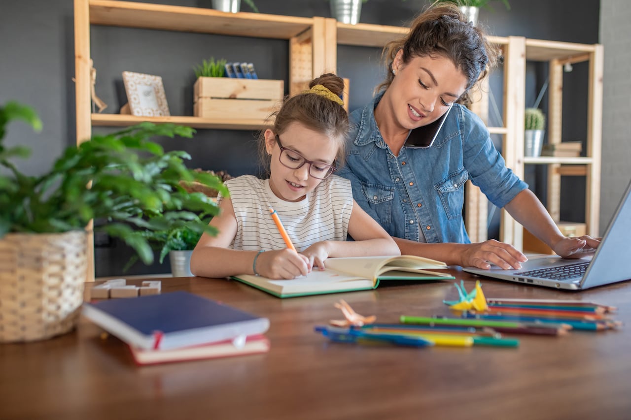 Madre e hija en casa. La niña está haciendo la tarea mientras su madre está trabajando en la computadora portátil y hablando por teléfono