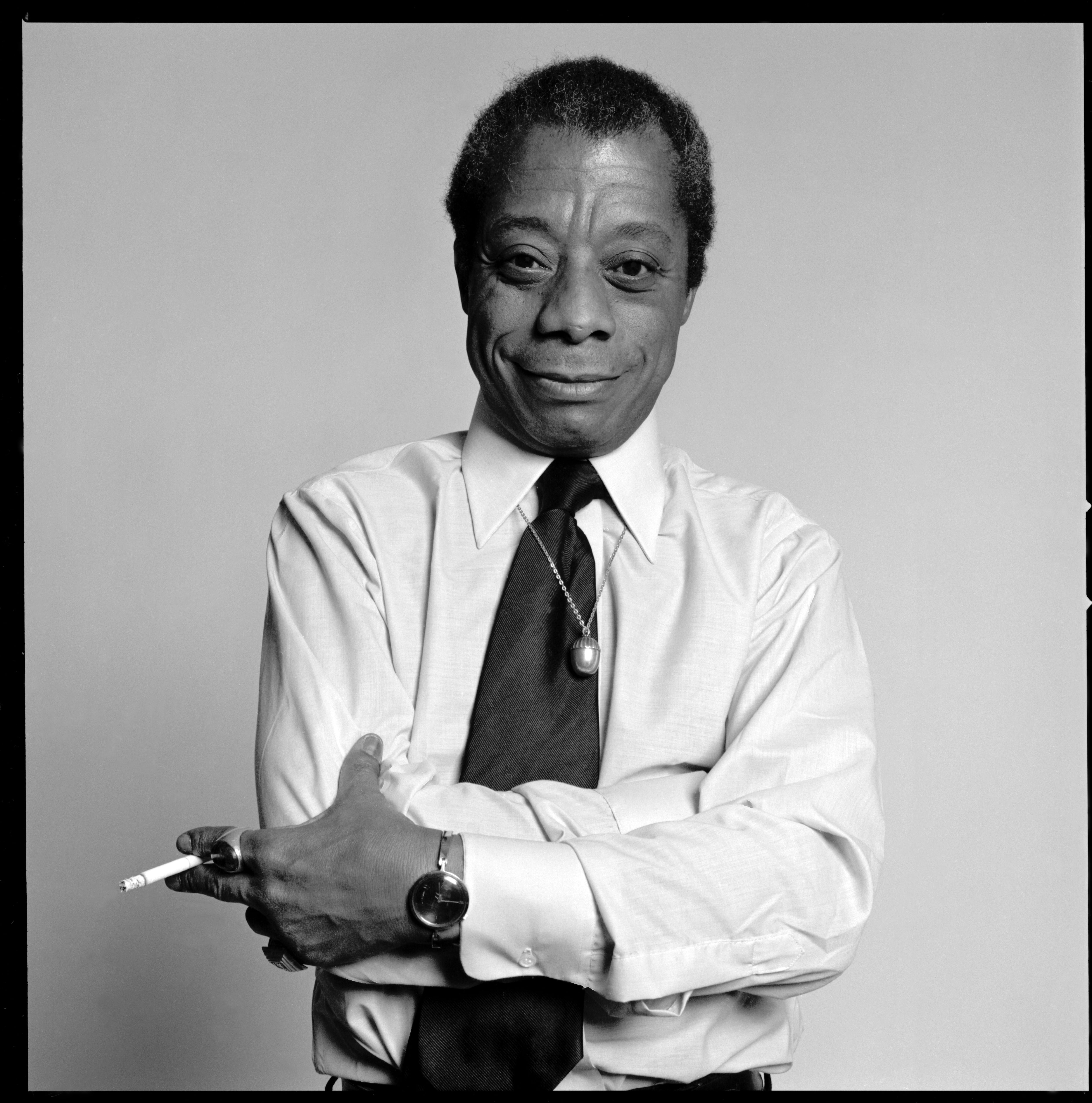 Fotografía en blanco y negro de James Baldwin sentado, con expresión contemplativa y vestimenta formal.