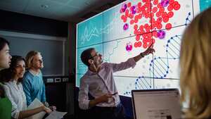 Grupo de profesionales de negocios en una habitación oscura parados frente a una gran pantalla de visualización de datos con información.