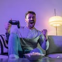 Young handsome man holding PlayStation joystick and cheerfully shouting . He sits on sofa in the living room and having great time with friends playing games online