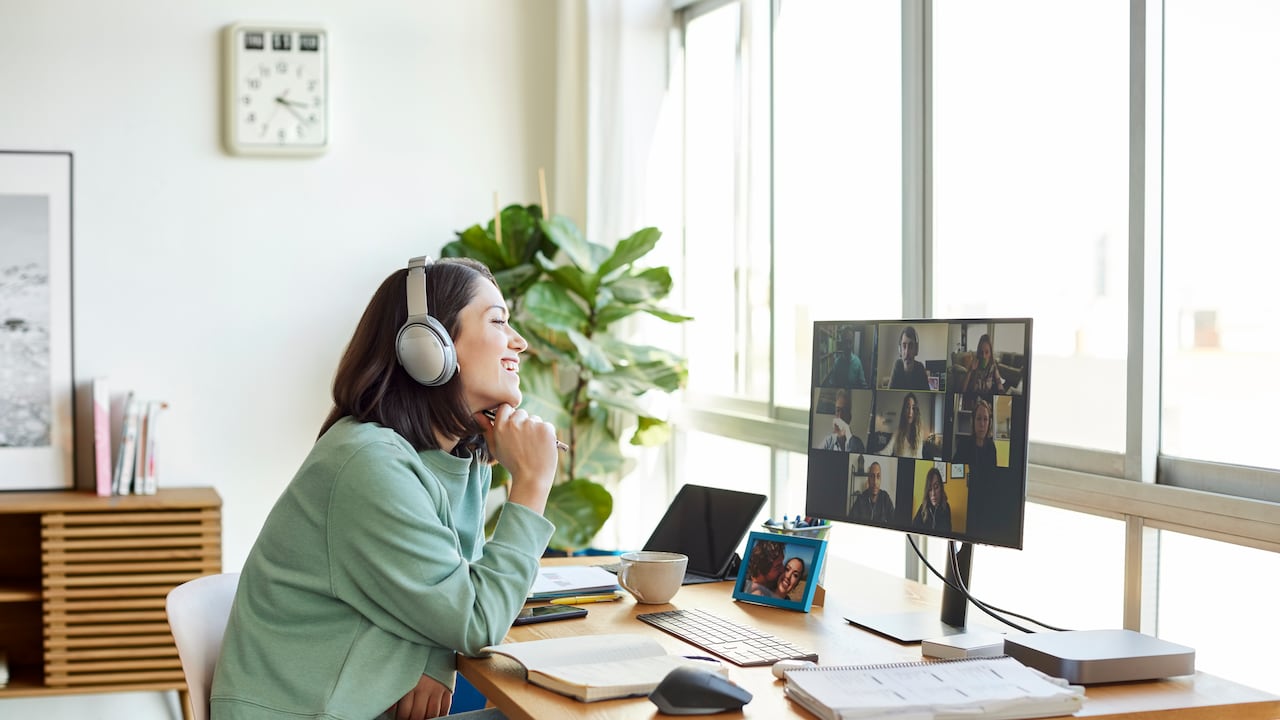 Smiling businesswoman discussing with colleagues through video call. Female professional is sitting in bright home office. She is listening through headphones.