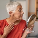 Hermosa anciana de pelo corto sosteniendo un espejo y aplicando crema facial en casa. Atractiva anciana de pelo gris mirándose en el espejo mientras se sienta en el sofá durante el día.