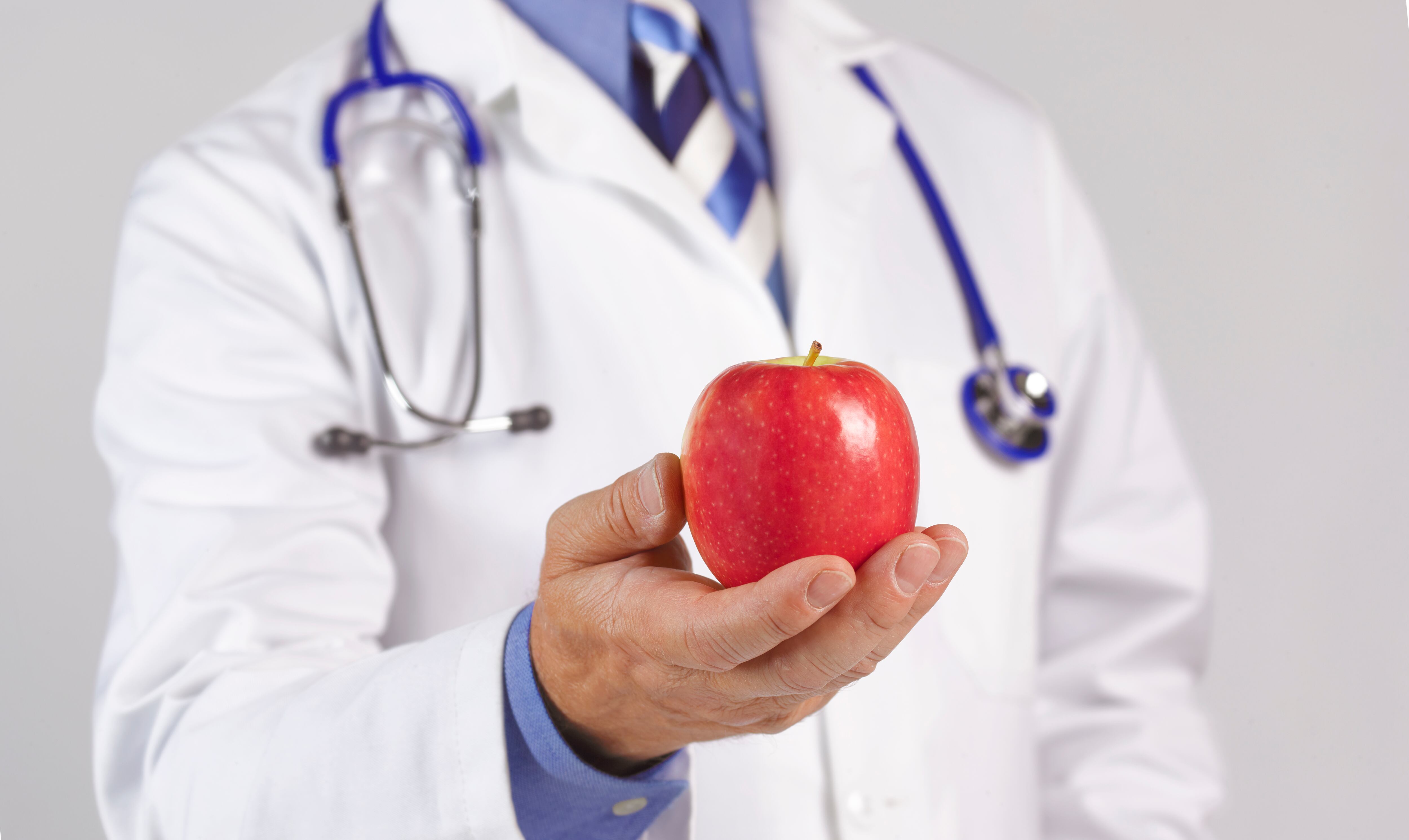 Doctor in white coat and stethoscope holding an apple to camera