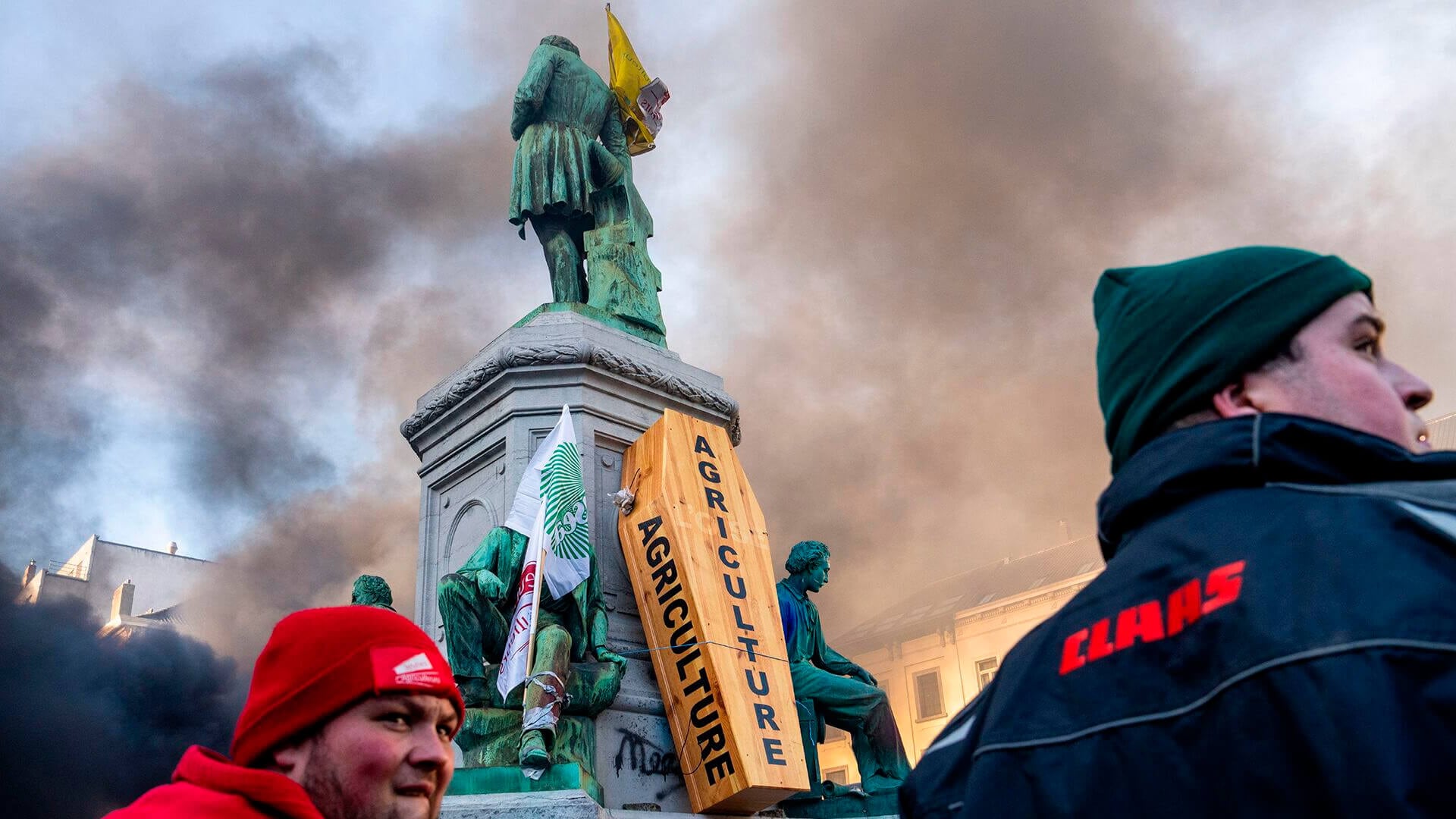 Hombres caminan junto a un ataúd de madera colocado sobre una base de estatuas durante una manifestación de agricultores europeos a las afueras de la Cumbre de la UE en Bruselas, el jueves 18 de diciembre de 2025.