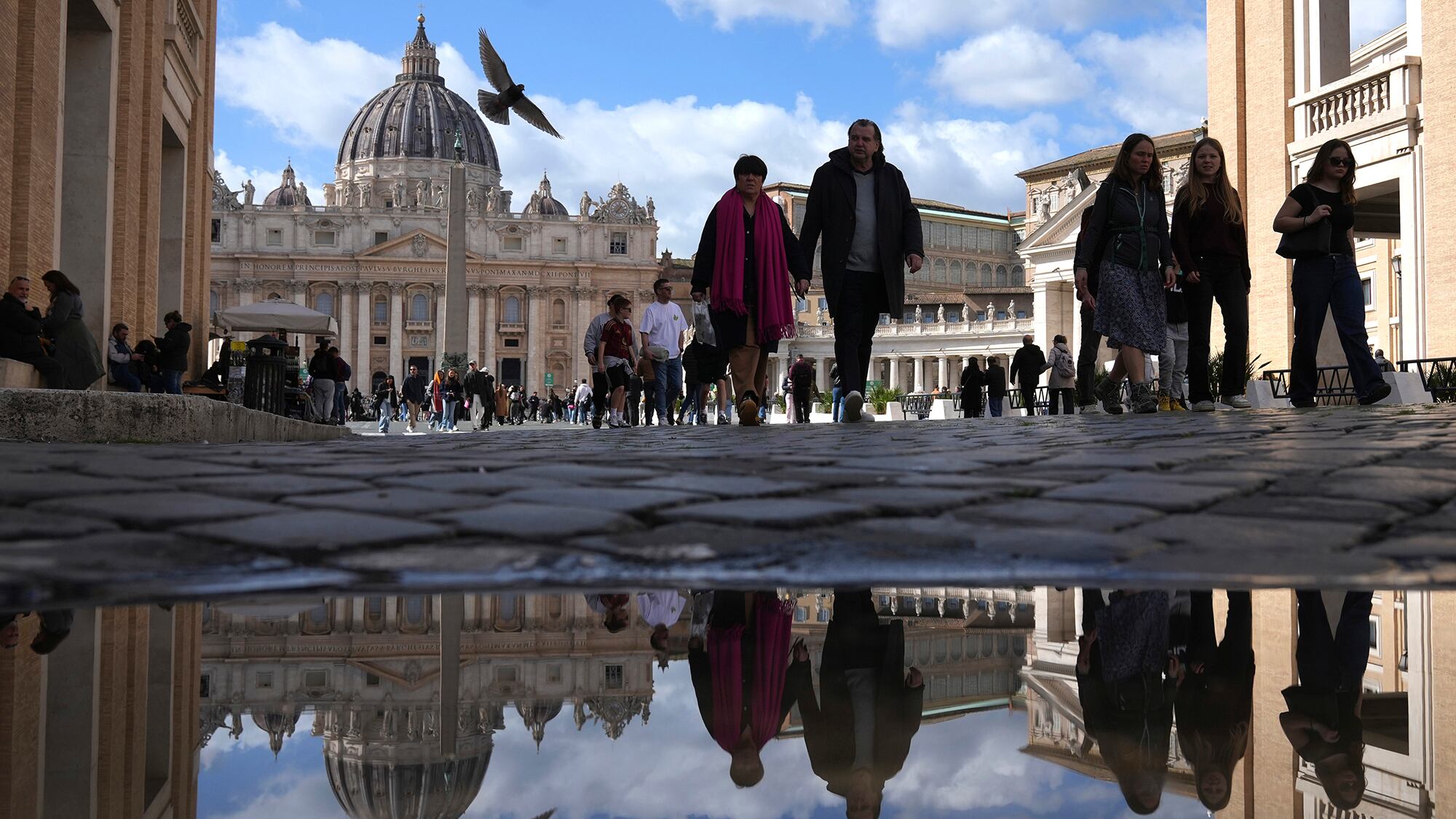 La gente se refleja en un charco mientras camina frente a la Plaza de San Pedro en el Vaticano. Jueves 27 de febrero de 2025.