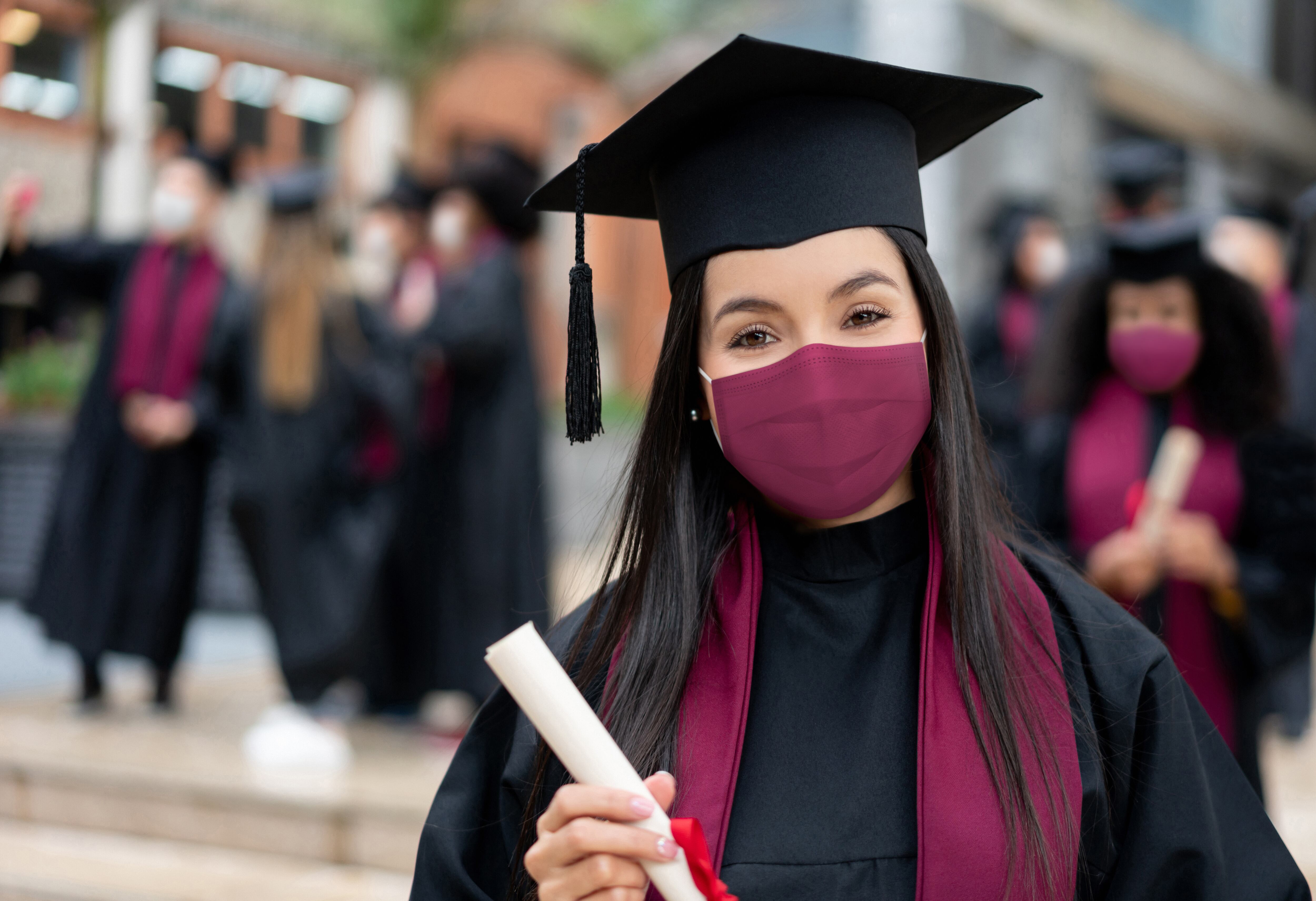 Retrato de una estudiante de posgrado feliz sosteniendo su diploma y con una mascarilla en su graduación durante la pandemia de COVID-19