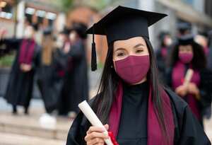 Retrato de una estudiante de posgrado feliz sosteniendo su diploma y con una mascarilla en su graduación durante la pandemia de COVID-19