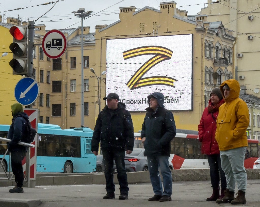 Los peatones cruzan una calle frente a una valla publicitaria que muestra el símbolo "Z" con los colores de la cinta de San Jorge y un lema que dice: "No nos damos por vencidos con nuestro pueblo", en apoyo de las fuerzas armadas rusas, en San Petersburgo, 7 de marzo de 2022 (Foto por AFP)