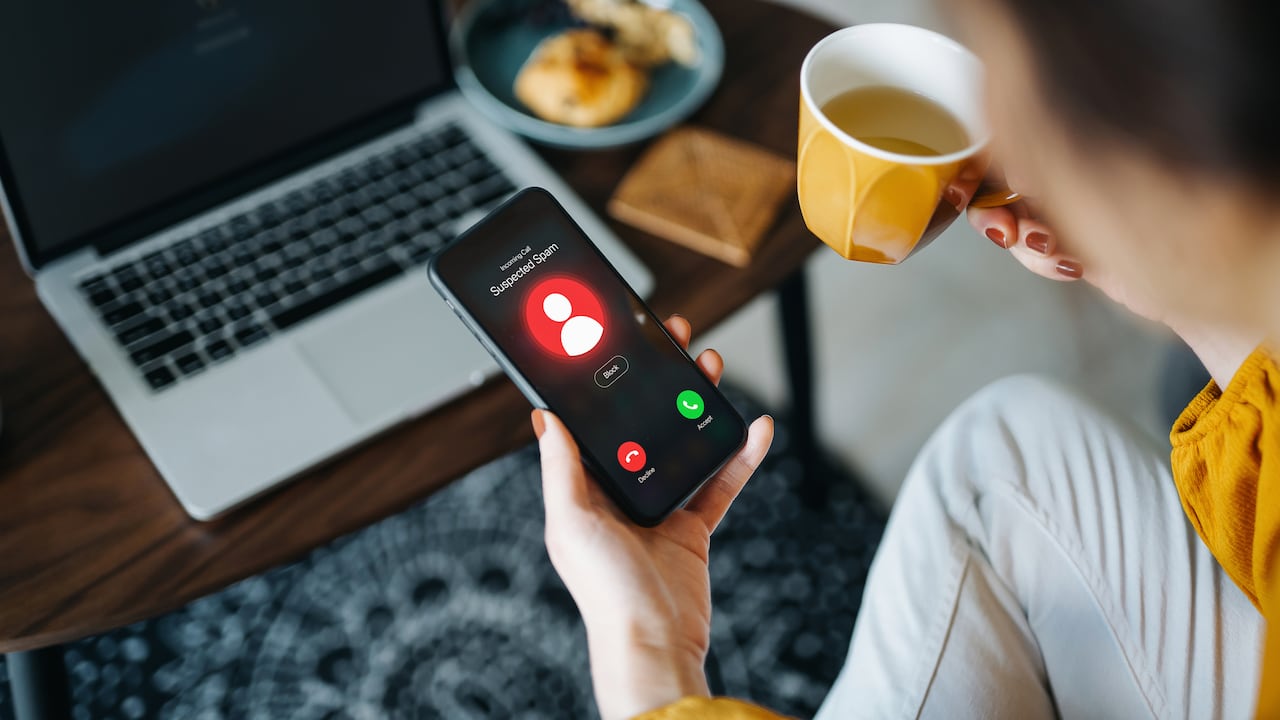 Over the shoulder view of young Asian woman receiving an incoming suspected call from unknown caller on her smartphone and rejecting the call at home. Device screen showing warning sign as detected by the network provider. Phone scam and fraud concept