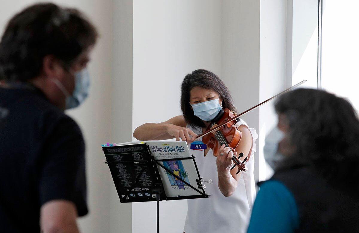 Un par de visitantes enmascarados miran cómo la enfermera Pam Guntrum, que trabaja en la educación del personal, toca un violín en un área de vestíbulo en Harborview Medical Center, en Seattle. Guntrum ha estado tocando su violín allí por años y hasta el momento nadie se lo ha impedido en el hospital. Los datos de los modelos de proyección de covid-19 muestran que la tasa de infección está aumentando en el estado de Washington, dijo el gobernador Jay Inslee, mientras instaba a la gente a seguir su enfoque medido para reabrir lentamente el estado de sus restricciones de estancia en casa. Foto: Elaine Thompson / AP.