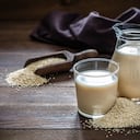 Vista frontal de una jarra y un vaso lleno de leche de quinua. El vaso y la jarra están en el lado derecho de la imagen sobre una mesa rústica de madera, al lado del vaso hay un montón de semillas de quinua. Esta imagen es parte de una serie de leches veganas.
