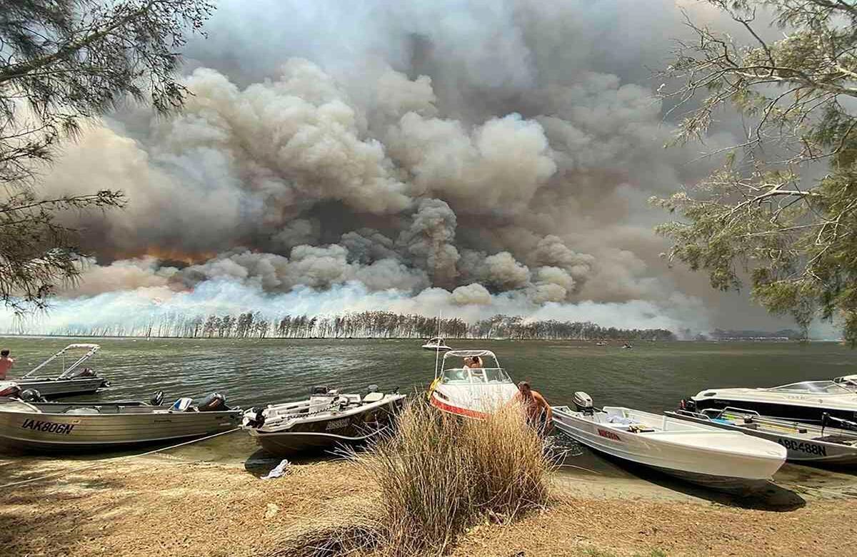Embarcaciones de diferentes tamaños son arrastradas a tierra mientras el humo y las llamas cubren gran parte del lago Conjola, popular destino turístico en Australia, el jueves 2 de enero del 2020. Miles de turistas huyeron de la costa este de Australia devastada por el fuego antes de que las condiciones se agravaran, cuando los militares comenzaron la labor de evacuar a las personas en la costa ubicada más al sur. Foto de Robert Oerlemans vía AP. 