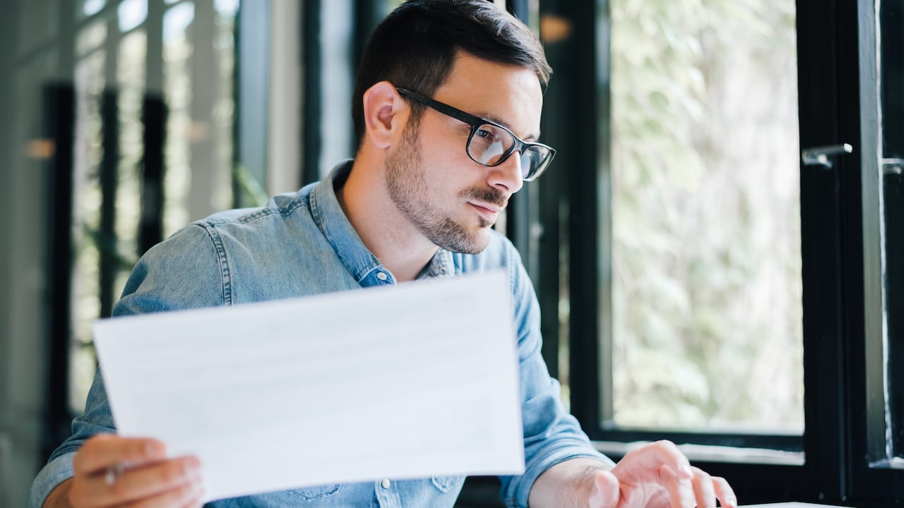 Serious pensive thoughtful focused young casual business accountant bookkeeper in office looking at and working with laptop and income tax return papers and documents