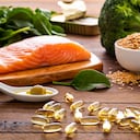 Front view of many fish oil capsules spilling out from the bottle surrounded by an assortment of food rich in omega-3 such as salmon, flax seeds, broccoli, sardines, spinach, olives and olive oil.